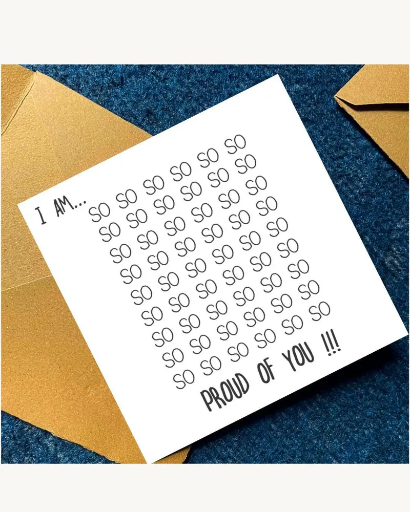 A white card with the text "I AM... SO SO SO SO SO SO... PROUD OF YOU !!!" is placed inside a gold e