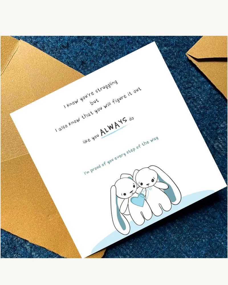 A white greeting card with a blue border and a cartoon illustration of two bunnies holding a blue heart.