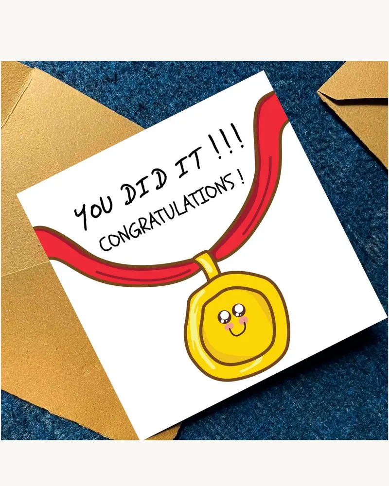 A square greeting card featuring a cartoon illustration of a gold medal on a red ribbon. The medal h